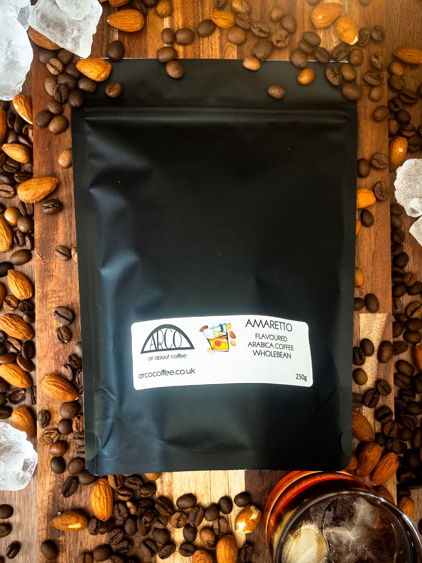 Bag of Arco Amaretto-flavoured Arabica wholebean coffee on a wooden surface, surrounded by coffee beans, almonds, ice cubes, and a glass of liqueur, reflecting its smooth roast infused with almond liqueur flavour