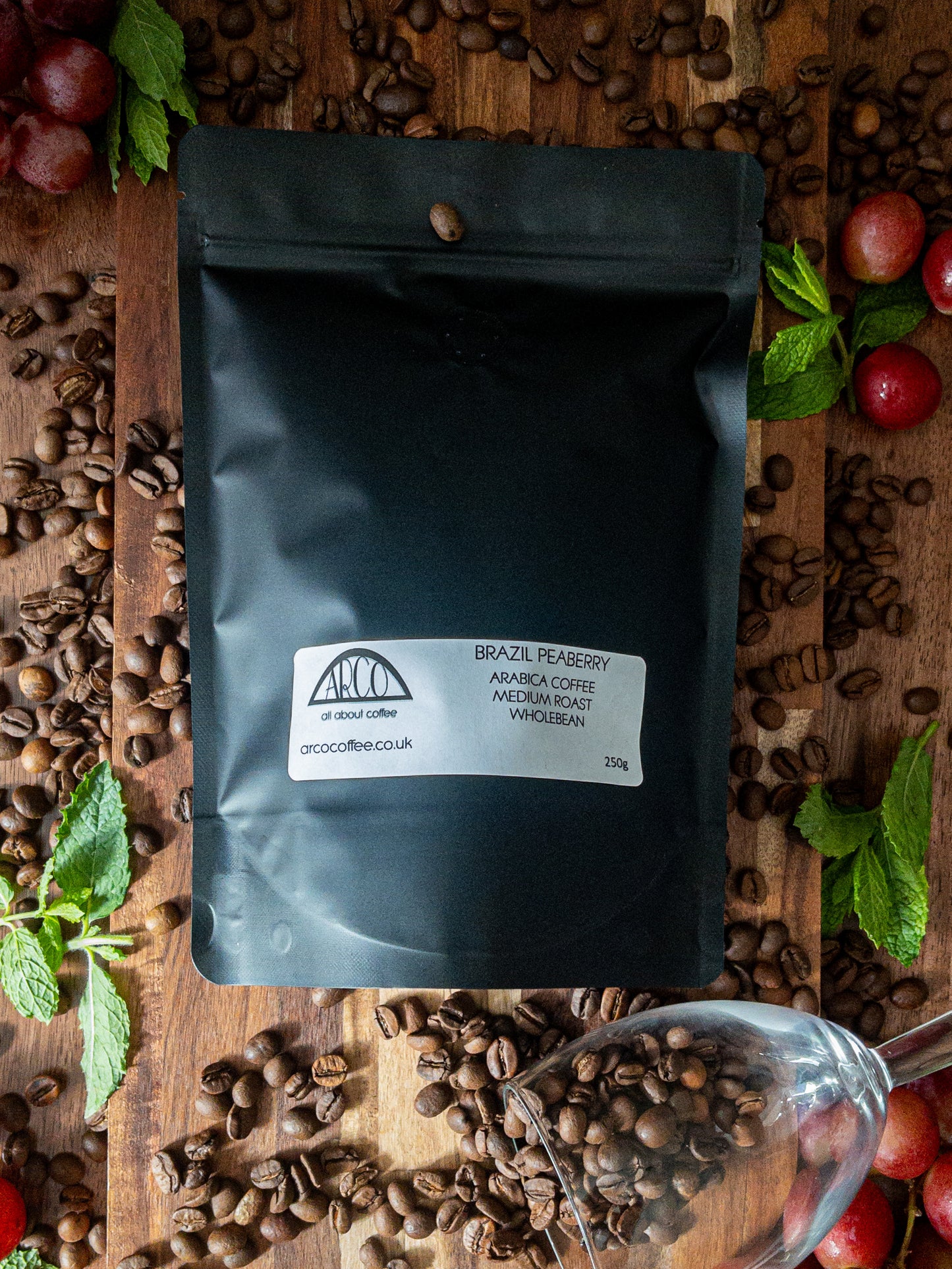 Bag of Arco Brazil Peaberry Arabica wholebean coffee on a wooden surface, surrounded by fresh grapes, and scattered coffee beans, representing its sharp, winey profile and fruity finish