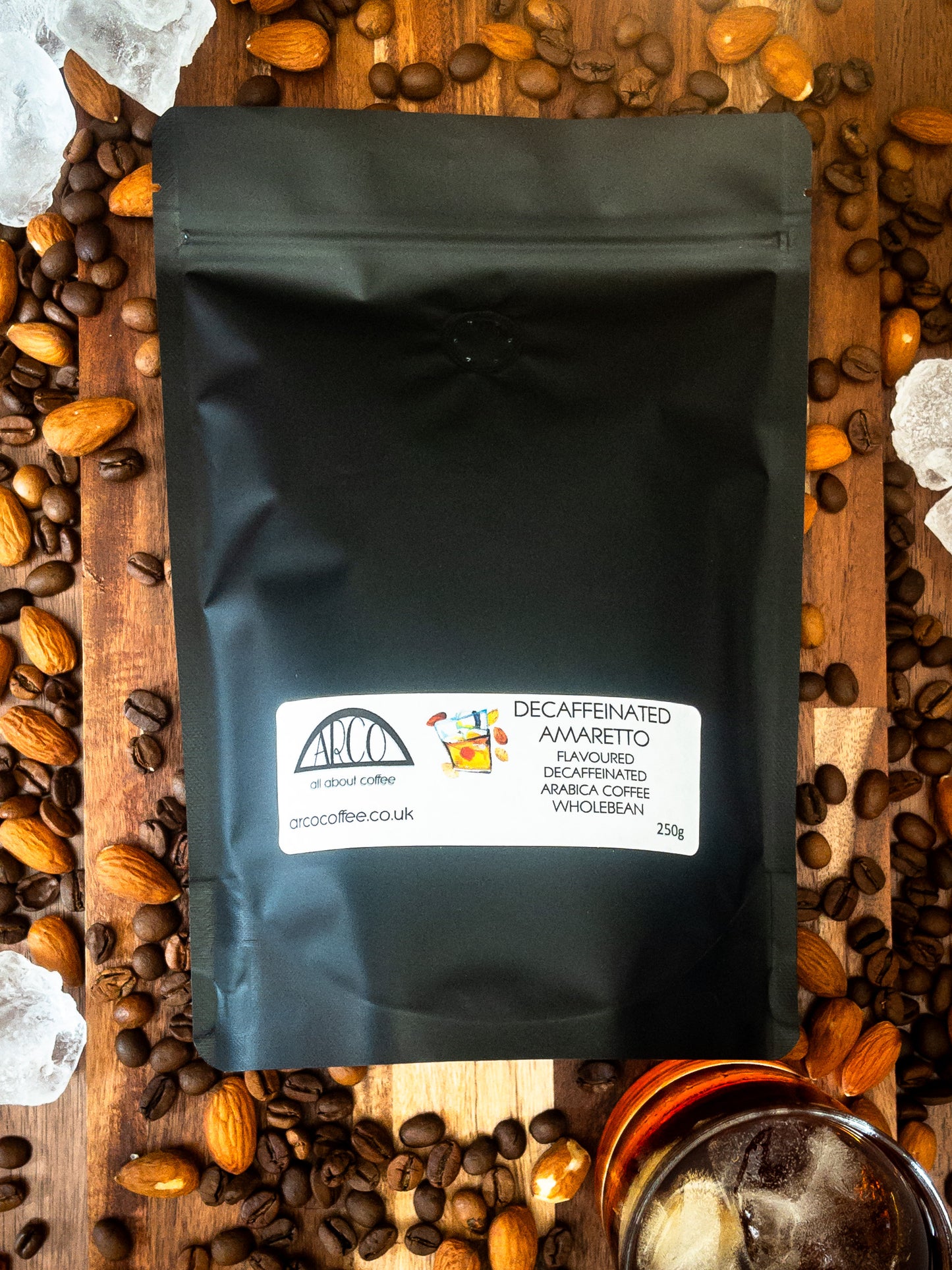 Bag of Arco decaffeinated Amaretto-flavoured Arabica wholebean coffee on a wooden surface, surrounded by coffee beans, almonds, ice cubes, and a glass of liqueur, reflecting its smooth roast infused with almond liqueur flavour
