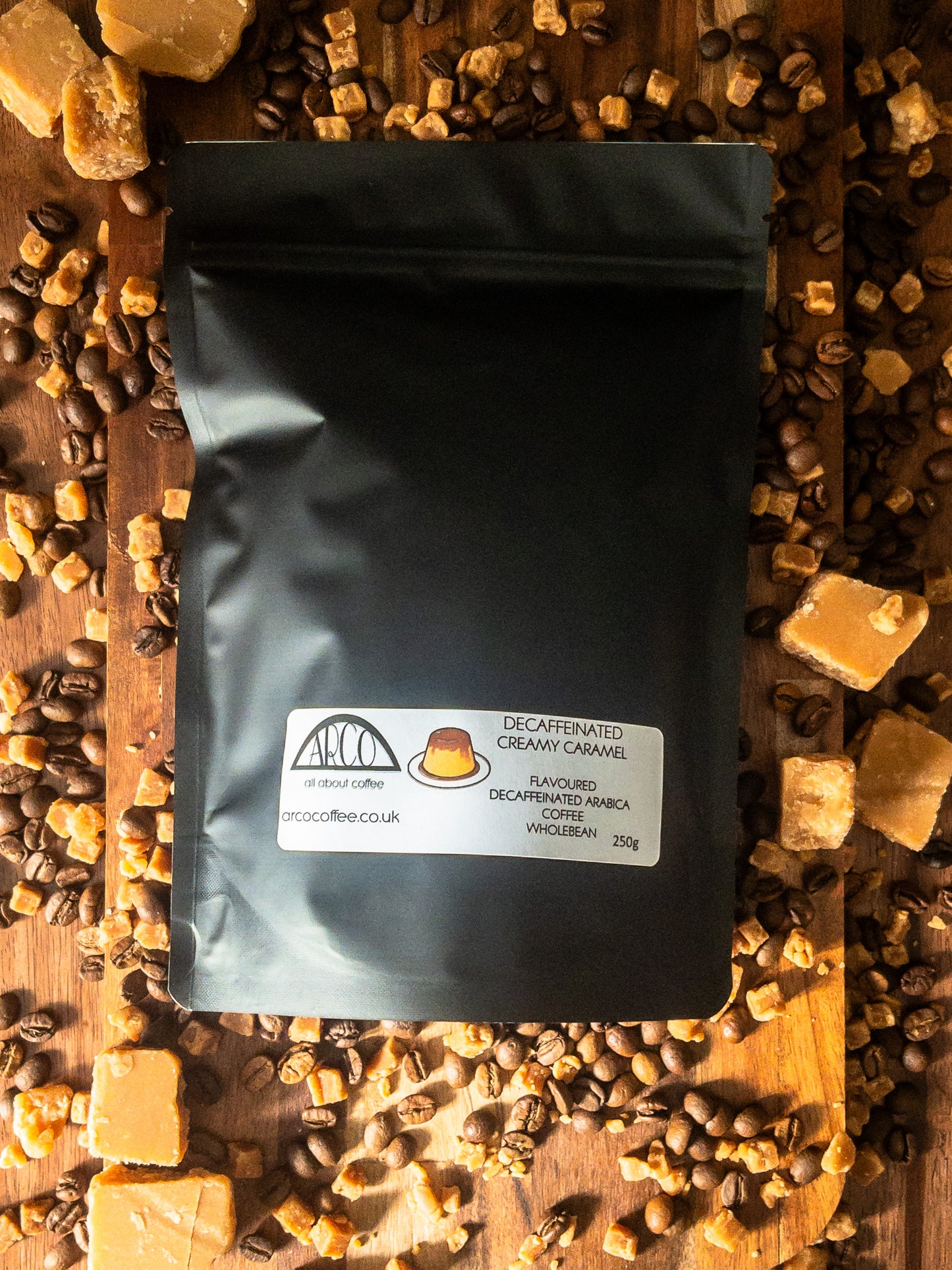 Bag of Arco decaffeinated Creamy Caramel Arabica wholebean coffee on a wooden surface, surrounded by coffee beans and caramel toffee pieces, capturing its smooth, classic flavour with rich caramel notes