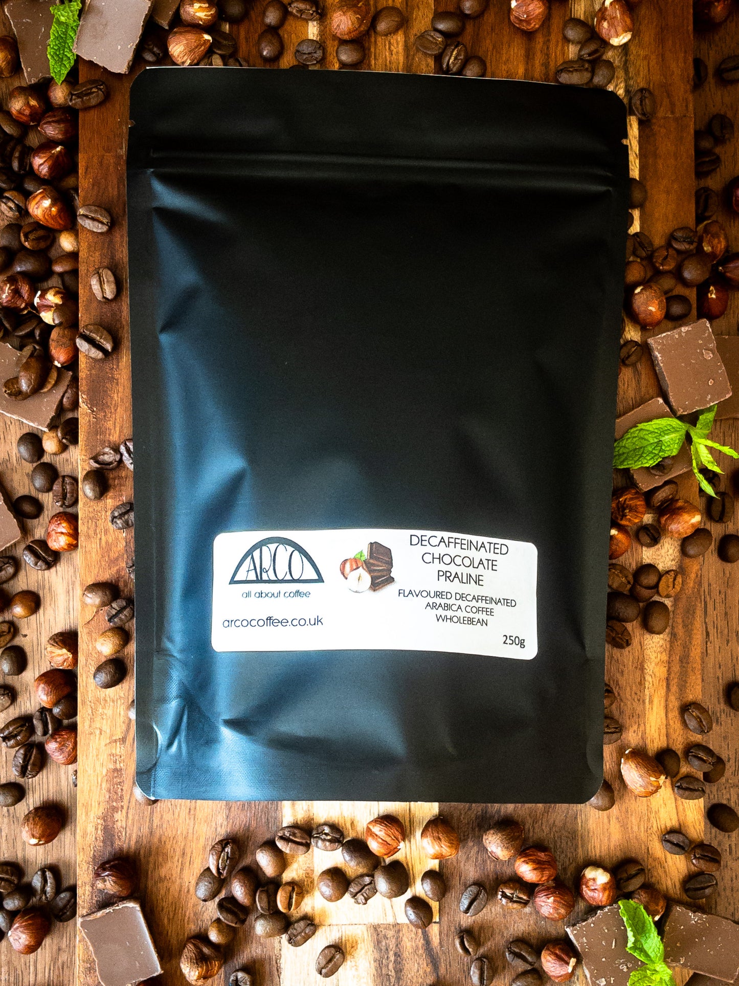 Bag of Arco decaffeinated Chocolate Praline Arabica wholebean coffee on a wooden surface, surrounded by coffee beans, chocolate pieces, and hazelnuts, highlighting its smooth medium roast with chocolate and nutty praline flavour