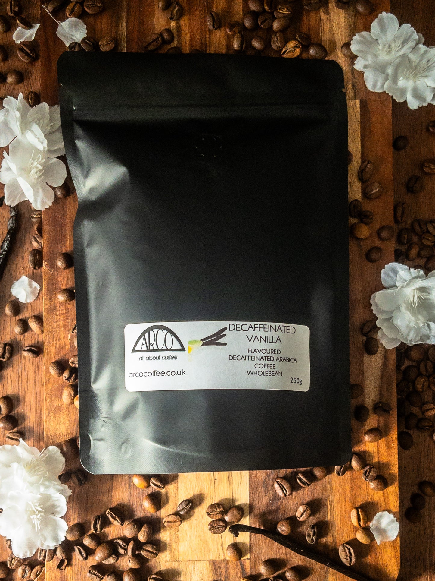 Bag of Arco decaffeinated vanilla-flavoured Arabica wholebean coffee on a wooden surface, surrounded by coffee beans, vanilla pods, and white blossoms, highlighting its smooth roast infused with Madagascan Bourbon vanilla
