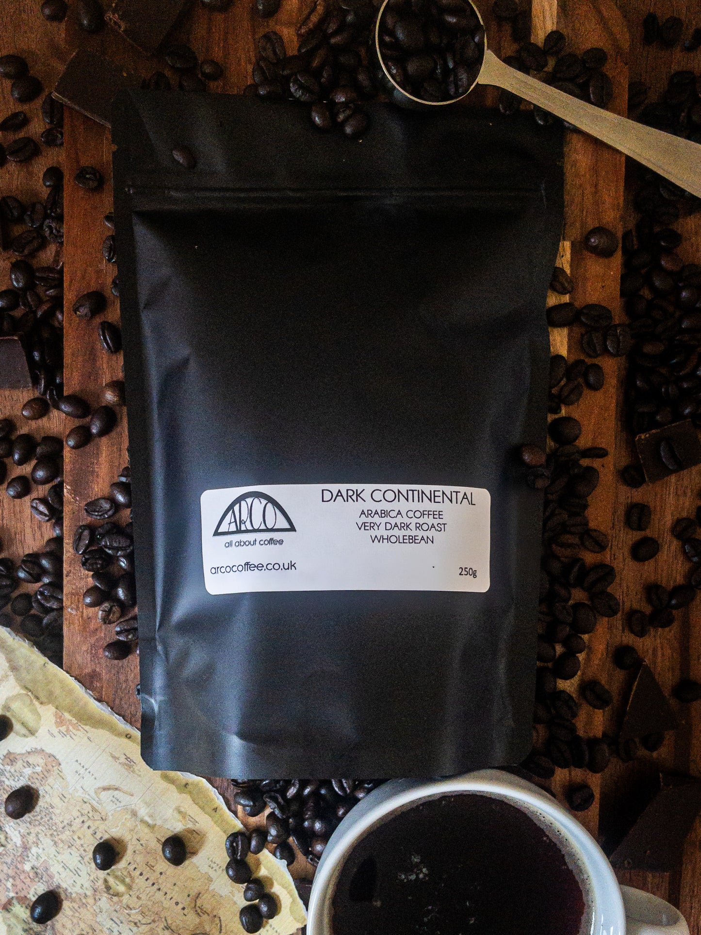 Bag of Arco Dark Continental Arabica wholebean coffee on a wooden surface, surrounded by dark roasted oily beans, a spoon, and a cup of black coffee, reflecting its rich, strong flavour and traditional very dark roast profile