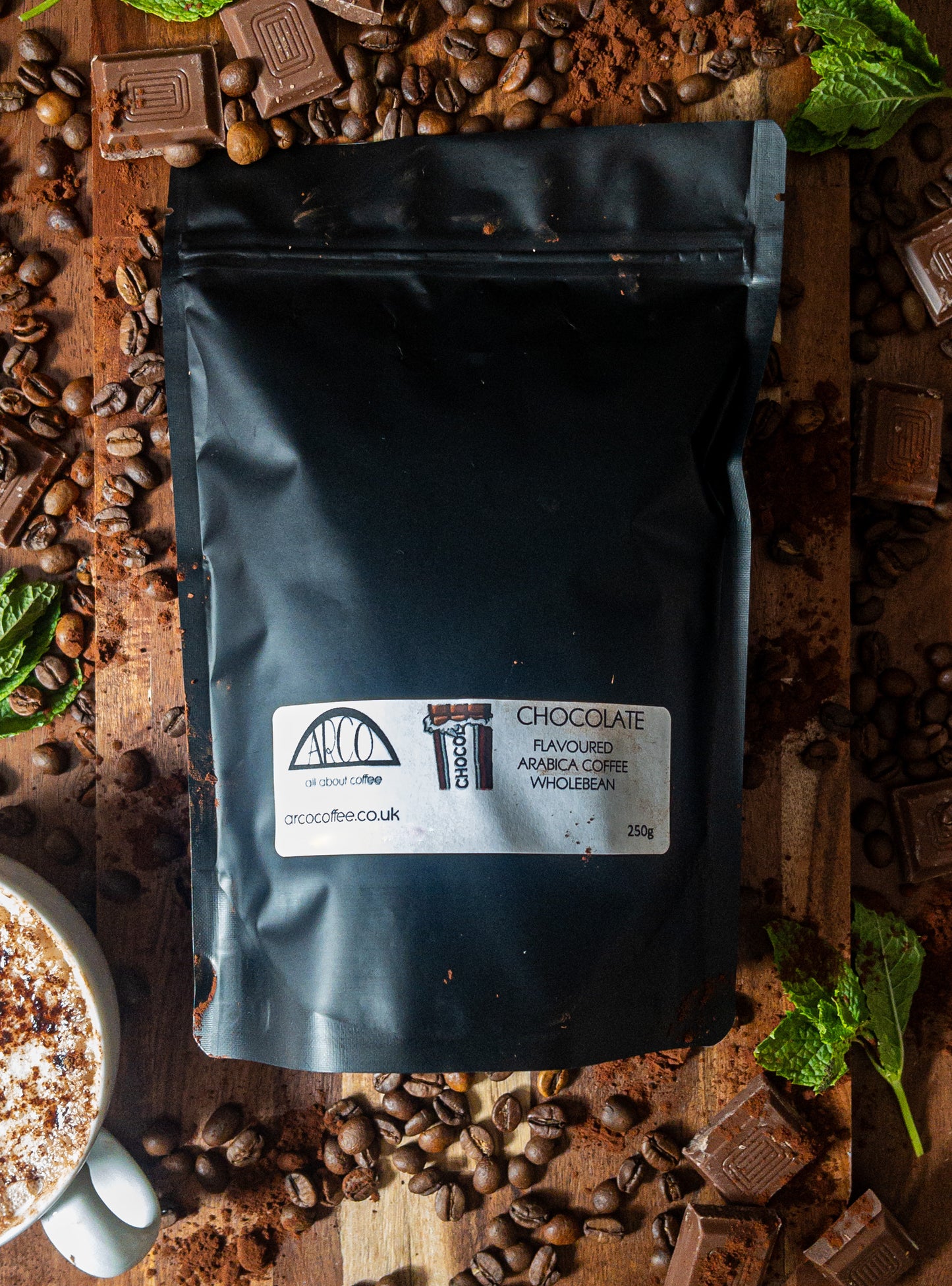 Bag of Arco chocolate-flavoured decaffeinated Arabica wholebean coffee on a wooden surface, surrounded by chocolate pieces, ground coffee, beans, and a foamy cappuccino, reflecting its smooth and naturally chocolate-infused profile