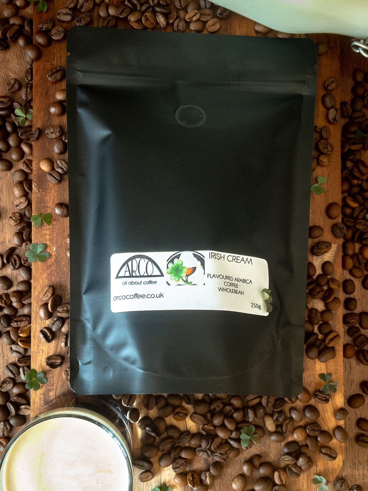 Bag of Arco Irish Cream-flavoured Arabica wholebean coffee on a wooden surface, surrounded by coffee beans, clover leaves, and a glass of cream coffee, reflecting its smooth roast infused with Irish whiskey and cream flavour