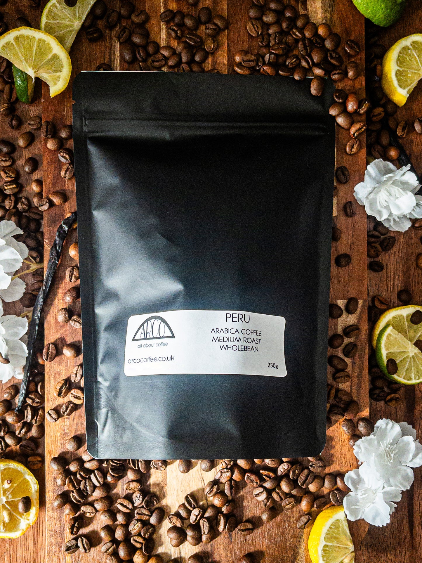 Bag of Arco Peru Arabica wholebean coffee on a wooden surface, surrounded by coffee beans, sliced lime, lemon, and white blossoms, reflecting its fruity and sweet flavour profile