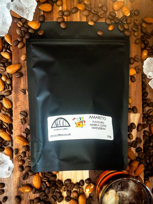 Bag of Arco Amaretto-flavoured Arabica wholebean coffee on a wooden surface, surrounded by coffee beans, almonds, ice cubes, and a glass of liqueur, reflecting its smooth roast infused with almond liqueur flavour