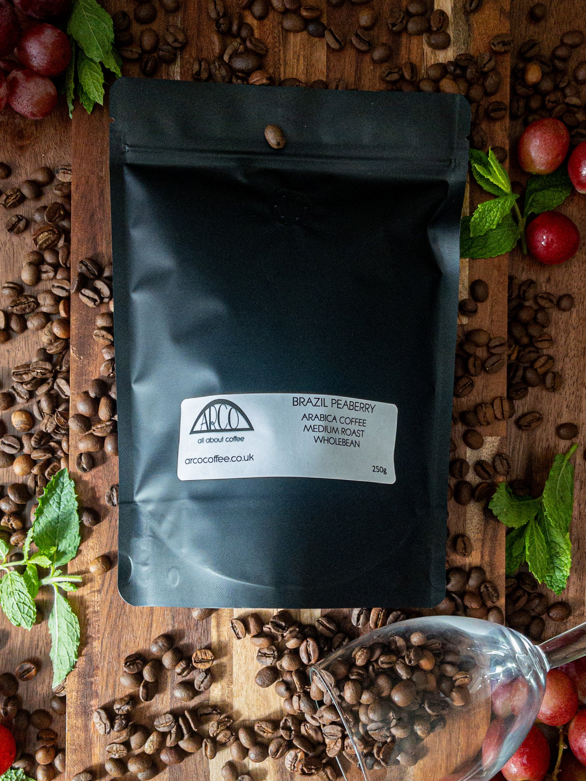 Bag of Arco Brazil Peaberry Arabica wholebean coffee on a wooden surface, surrounded by fresh grapes, and scattered coffee beans, representing its sharp, winey profile and fruity finish