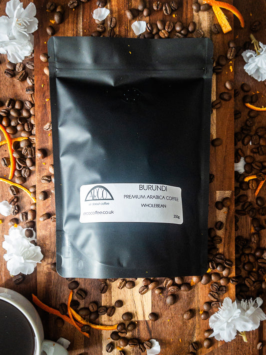 Bag of Arco Burundi premium Arabica wholebean coffee on a wooden surface, surrounded by coffee beans, white blossoms, and orange peel strips, reflecting its delicate flavour with floral notes of jasmine and orange blossom