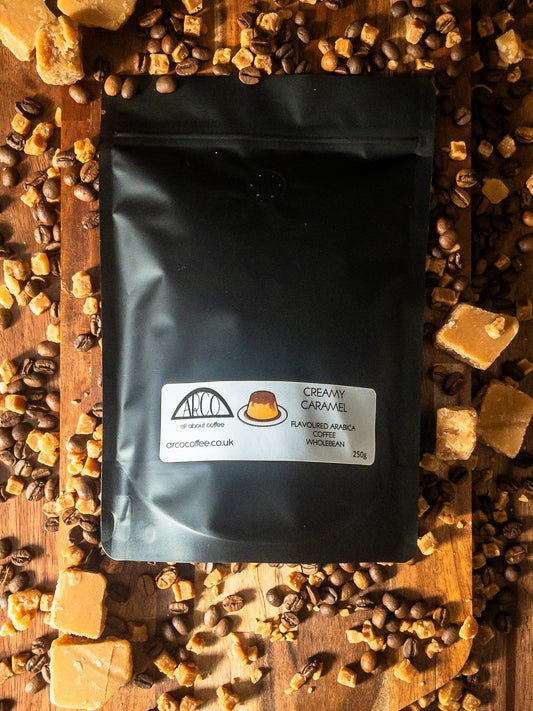 Bag of Arco Creamy Caramel Arabica wholebean coffee on a wooden surface, surrounded by coffee beans and caramel toffee pieces, capturing its smooth, classic flavour with rich caramel notes