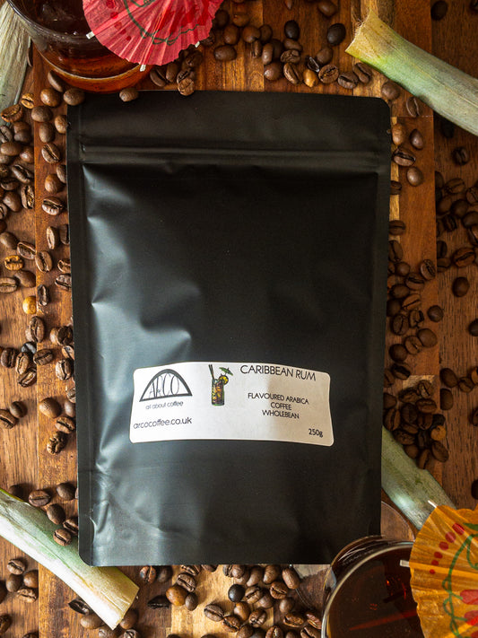 Bag of Arco Caribbean Rum-flavoured Arabica wholebean coffee on a wooden surface, surrounded by coffee beans, rum glasses, and tropical drink decorations, reflecting its smooth roast infused with rich Caribbean rum flavour