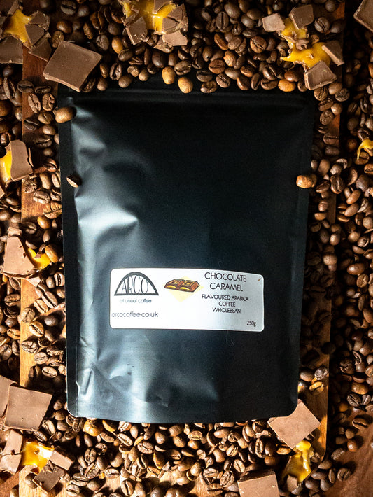 Bag of Arco Chocolate Caramel-flavoured Arabica wholebean coffee on a wooden surface, surrounded by coffee beans, chocolate chunks, and caramel pieces, highlighting its smooth medium roast with sweet, rich flavour notes