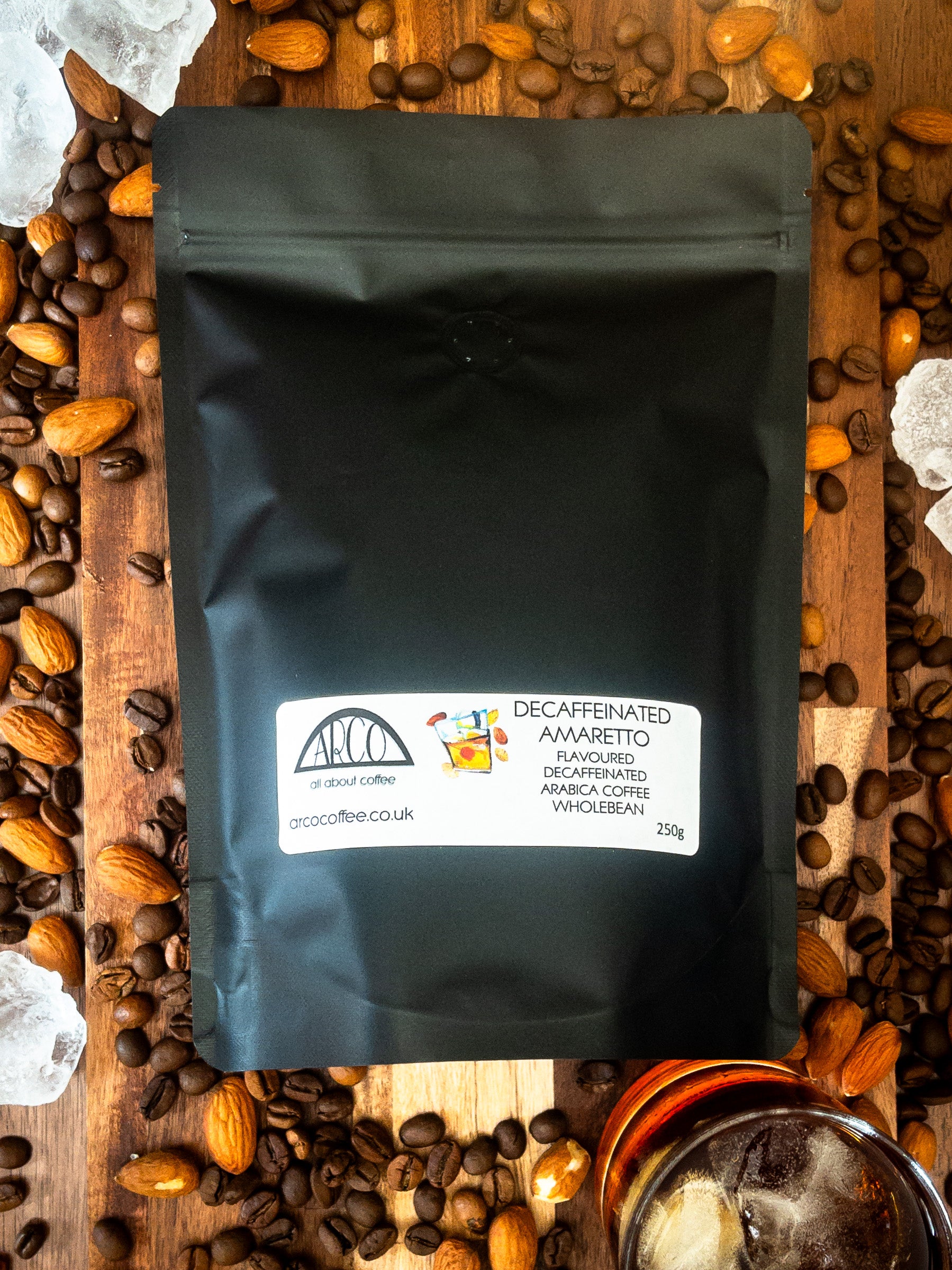 Bag of Arco decaffeinated Amaretto-flavoured Arabica wholebean coffee on a wooden surface, surrounded by coffee beans, almonds, ice cubes, and a glass of liqueur, reflecting its smooth roast infused with almond liqueur flavour