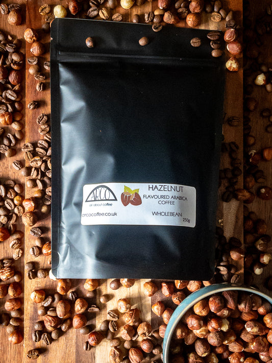 Bag of Arco hazelnut-flavoured Arabica wholebean coffee on a wooden surface, surrounded by coffee beans and scattered hazelnuts, reflecting its smooth roast and classic nutty flavour
