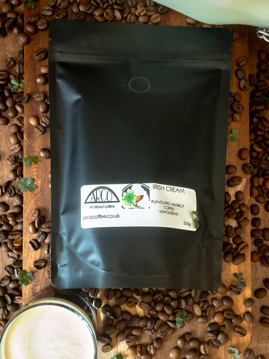 Bag of Arco Irish Cream-flavoured Arabica wholebean coffee on a wooden surface, surrounded by coffee beans, clover leaves, and a glass of cream coffee, reflecting its smooth roast infused with Irish whiskey and cream flavour