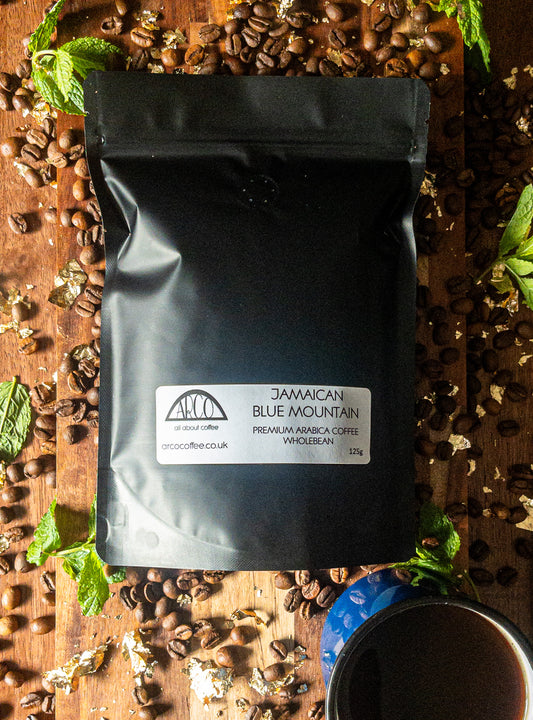 Bag of Arco Jamaican Blue Mountain premium Arabica wholebean coffee on a wooden surface, surrounded by coffee beans, gold flakes, and a cup of black coffee, reflecting its rare and world-renowned quality