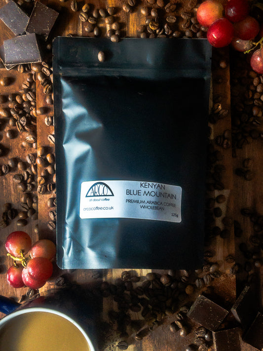 Bag of Arco Kenyan Blue Mountain premium Arabica wholebean coffee on a wooden surface, surrounded by coffee beans, dark chocolate squares, red grapes, and a cup of coffee, reflecting its rich body, cocoa notes, and all-day drinkability