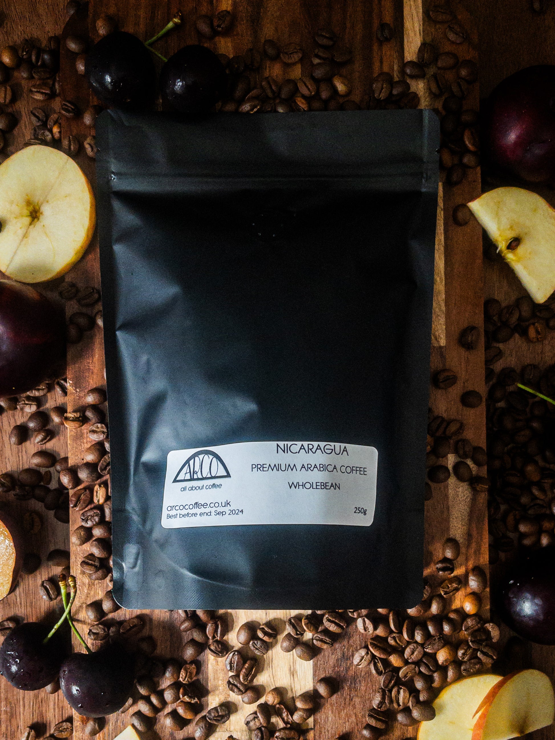 Bag of Arco Nicaragua premium Arabica wholebean coffee on a wooden surface, surrounded by coffee beans, sliced apples, cherries, and dark plums to represent its fruity and spicy tasting notes.