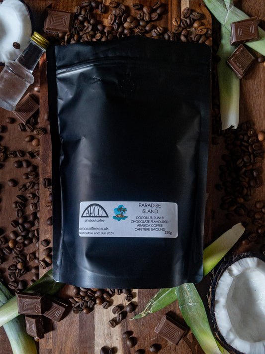 Bag of Arco Paradise Island chocolate, rum, and coconut-flavoured Arabica wholebean coffee on a wooden surface, surrounded by coffee beans, coconut pieces, chocolate squares, and tropical fruit, reflecting its indulgent tropical flavour profile