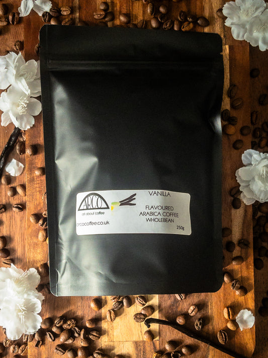 Bag of Arco vanilla-flavoured Arabica wholebean coffee on a wooden surface, surrounded by coffee beans, vanilla pods, and white blossoms, highlighting its smooth roast infused with Madagascan Bourbon vanilla