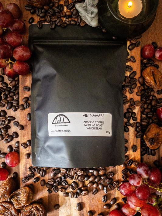 Bag of Arco Vietnamese Arabica medium roast wholebean coffee on a wooden surface, surrounded by coffee beans, red grapes, dried figs, and a lit candle, evoking a rich, full-bodied and subtly fruity character